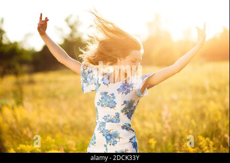 Belle tendre mignonne heureuse jeune fille jette ses mains belle blonde dans un champ de fleurs dans une robe multicolore sur fond de lumière de coucher de soleil Banque D'Images