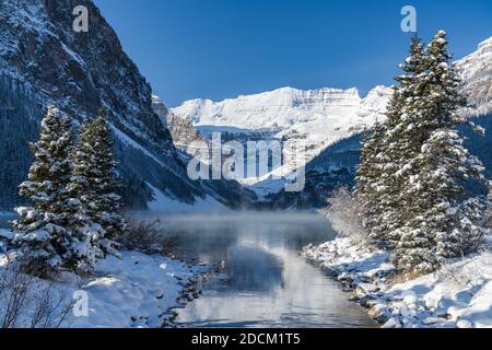 Lac Louise en début d'hiver, jour ensoleillé le matin. Brume flottant sur la surface turquoise de l'eau. Ciel bleu clair, montagnes enneigées en arrière-plan Banque D'Images