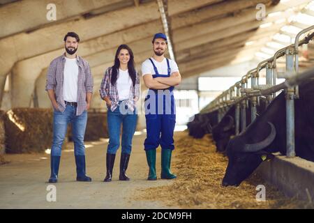 Groupe de jeunes éleveurs de bovins heureux debout dans un étable de bétail et souriant Banque D'Images