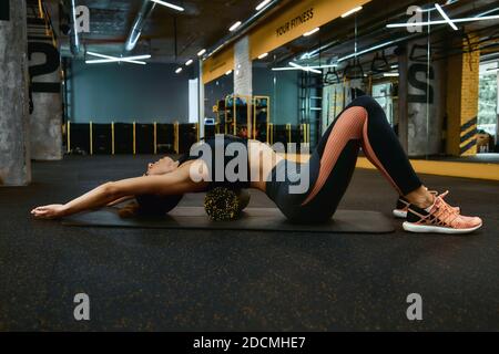 Détente totale après l'entraînement. Pleine longueur d'une jeune femme sportive dans les vêtements de sport s'exerçant avec le rouleau en mousse, allongé sur le tapis de yoga à la salle de gym et faisant des exercices d'étirement. Le sport et un mode de vie sain Banque D'Images