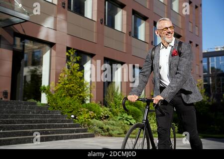 Trajets quotidiens. Homme d'affaires d'âge moyen, élégant et réussi, en lunettes de soleil souriant tout en marchant avec son vélo pour travailler et en regardant loin dans la rue urbaine Banque D'Images