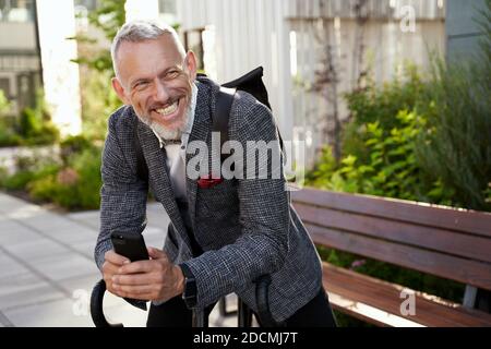 Joyeux homme d'affaires d'âge moyen en costume élégant souriant tout en tenant son smartphone, debout avec un vélo à l'extérieur dans la journée Banque D'Images