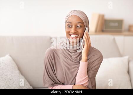 Femme noire souriante dans le hijab parlant sur un smartphone à la maison Banque D'Images