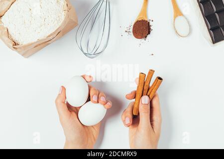 Mains de femmes tenant des oeufs et des bâtons de cannelle à côté des ingrédients de cuisson, de la farine, de la poudre de cacao, du chocolat et du sucre sur fond blanc, ponte plate. Banque D'Images