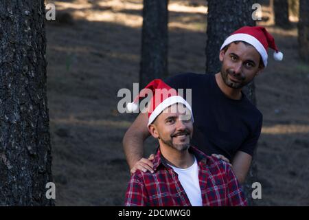 Couple gay souriant avec chapeaux de Noël rouges posé dans le parc forestier. Des hommes heureux célébrant la saison des vacances d'hiver dans la nature en plein air Banque D'Images