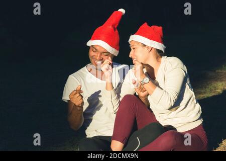 Jeunes couples Red chapeaux chantant des chants de Noël assis sur le sol sur la forte lumière du soleil à l'extérieur. Homme et femme brésiliens appréciant les vacances d'hiver Banque D'Images