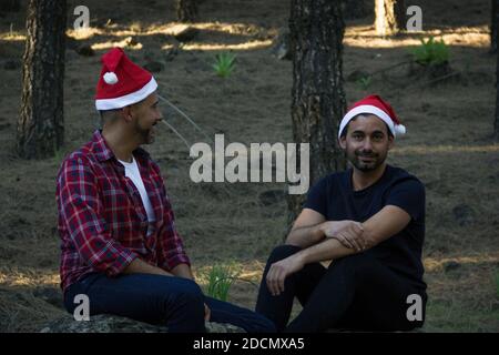 Jeune couple gay avec des chapeaux de Noël rouges assis sur le parc forestier en pin. Deux hommes posant dans la saison des vacances d'hiver en plein air dans la nature Banque D'Images