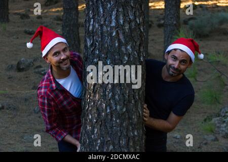 Portrait de couple d'amis mâles avec chapeaux de Noël rouges sortant de l'arbre dans le parc forestier. Hommes souriants prêts pour les vacances d'hiver en plein air Banque D'Images