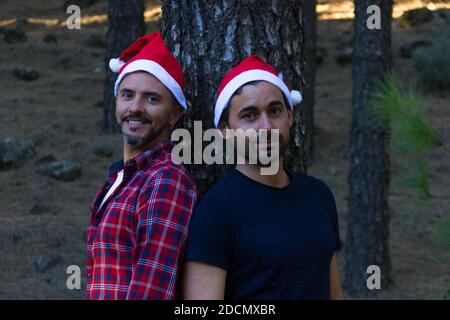 Deux hommes avec des chapeaux de Noël rouges pendu tronc de pin dans le parc forestier. Couple gay souriant posant dans la saison des vacances d'hiver dans la nature Banque D'Images