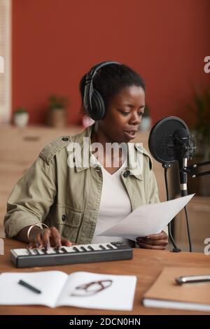 Portrait vertical de la jeune femme afro-américaine qui compose de la musique et chante pour un microphone dans un studio d'enregistrement à domicile Banque D'Images