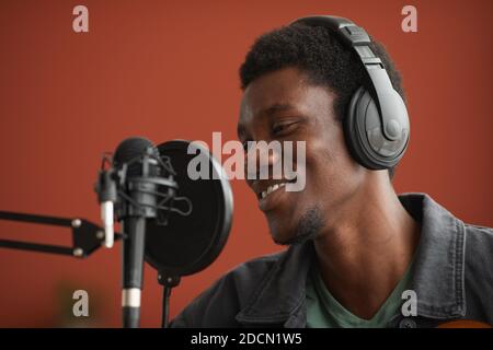 Gros plan d'un homme afro-américain souriant chantant au microphone sur fond rouge dans un studio d'enregistrement, espace de copie Banque D'Images