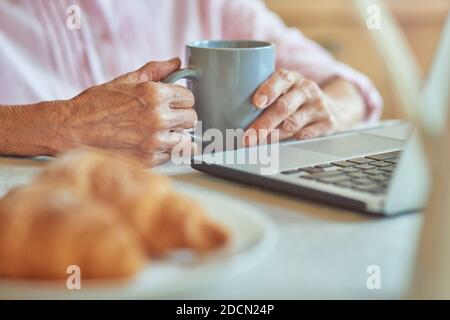 Photo rognée d'une femme âgée tenant un café et utilisant un ordinateur portable pendant le petit déjeuner le matin. Concept de style de vie domestique Banque D'Images