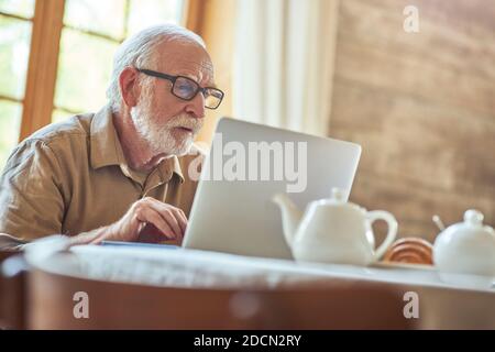 Un retraité de sexe masculin dans des lunettes regarde attentivement l'écran d'un ordinateur portable tout en travaillant à la maison. Concept de style de vie Banque D'Images