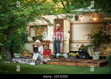 Homme de grande famille souriant offrant un croissant à sa femme qui utilise une tablette numérique près de leur camping-car. Concept de voyage Banque D'Images