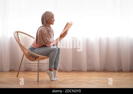 Femme afro-américaine attirante dans hijab lisant un livre fascinant dans un fauteuil confortable à la maison, espace vide Banque D'Images