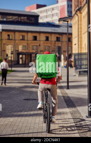 Vue arrière d'un coursier avec sac thermo à vélo le long de la rue carrelée sur une journée ensoleillée, plein plan vertical. Concept de livraison de nourriture Banque D'Images