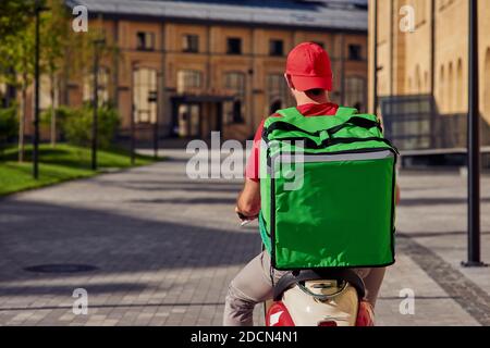 Vue arrière d'un messager mâle avec sac thermo à vélo le long d'une belle rue ensoleillée. Concept de livraison de nourriture Banque D'Images