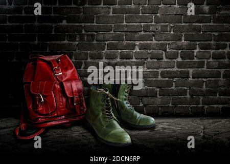 Bottes vert vintage et sac à dos rouge en cuir véritable sur fond de mur de briques sombres. Banque D'Images