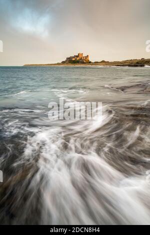 Une vague se brise au-dessus des Harkness Rocks à marée haute tandis que le château de Bamburgh est éclairé par la lumière basse de l'heure d'or à travers la baie. Banque D'Images