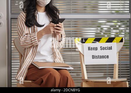 Portrait rogné d'une jeune femme portant un masque et utilisant un smartphone en attendant dans la file d'attente au bureau avec le signe garder social distance, espace de copie Banque D'Images