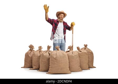 Jeune fermier barbu avec une pelle debout derrière des sacs de toile de jute et en agitant la caméra isolée sur fond blanc Banque D'Images