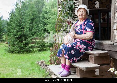 Grand-mère senior chapeau habillé et lunettes embrassant chat sphynx dans les mains tout en étant assis sur les escaliers en bois de porche, maison en bois dans la campagne, copyspac Banque D'Images