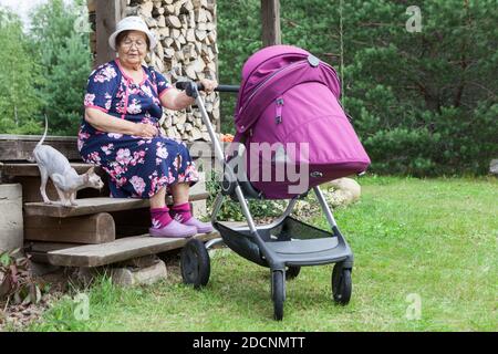 Bonne femme d'âge de retraite prend soin de dormir grand petit-enfant dans bébé pram, assis avec chat sphynx sur une véranda en bois au soleil Banque D'Images