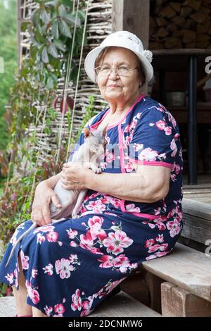 Femme âgée avec son portrait de chat sphynx, lunettes et housecavoine, assise sur le porche de maison de campagne en bois Banque D'Images