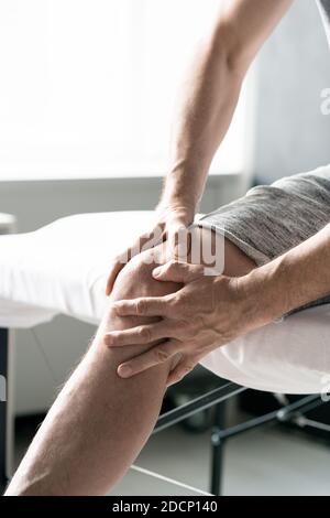 Les mains d'un homme mûr massent le genou droit en étant assis sur une table médicale Banque D'Images
