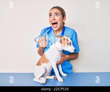 Jeune belle blonde vétérinaire femme mettant le vaccin au chien de chiot en colère et fou criant frustré et furieux, criant avec la colère regardant vers le haut. Banque D'Images