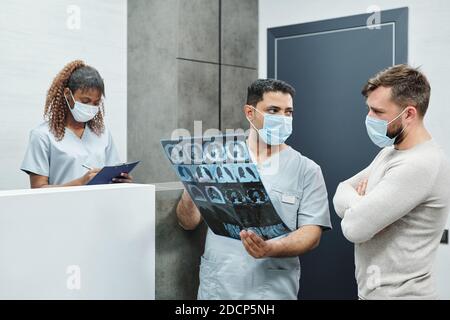 Radiologue masculin sérieux en uniforme et masque montrant une image radiologique au patient Banque D'Images