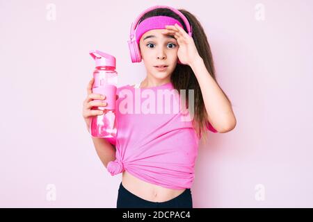 Adorable enfant hispanique fille portant des vêtements de gym et utilisant des écouteurs tenant une bouteille d'eau stressée et frustrée avec la main sur la tête, surpris et ang Banque D'Images