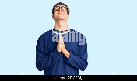 Jeune homme beau portant des vêtements et des lunettes décontractés mendiant et priant avec les mains avec l'expression de l'espoir sur le visage très émotionnel et inquiet. Banque D'Images