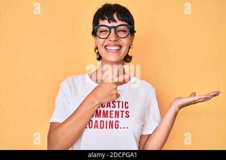 Belle femme brunettte portant des commentaires sarcastique chargement t-shirt étonné et sourire à l'appareil photo tout en présentant avec la main et en pointant avec ailette Banque D'Images