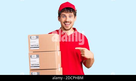 Jeune homme beau avec des cheveux bouclés tenant le paquet de livraison souriant contenchez-vous de pointer avec votre main et votre doigt Banque D'Images