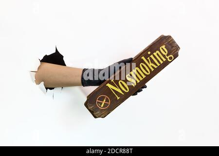 Une main femelle dans un gant noir en latex dans un trou déchiré sur un fond blanc tient un panneau en bois non-fumeurs. Mesures de quarantaine pour prévenir la propagation de Banque D'Images