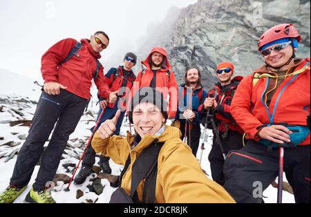 Jeune homme prenant des photos avec des amis randonneurs pendant les voyages dans les montagnes d'hiver. Groupe de voyageurs masculins regardant l'appareil photo et souriant, montrant les pouces vers le haut. Concept de voyage, de randonnée et de photographie. Banque D'Images