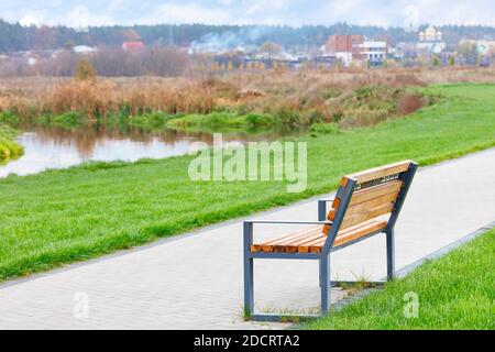 Un banc en bois se dresse sur un sentier de randonnée le long d'une belle pelouse verte sur le remblai de la rivière avec un fond flou d'un paysage rural. Banque D'Images