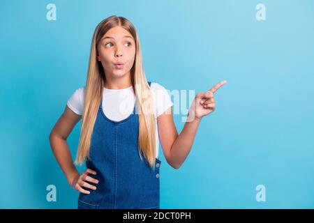 Photo d'adolescent surprise blonde longue fille point look vide space wear blanc t-shirt robe denim isolée sur la couleur bleue arrière-plan Banque D'Images