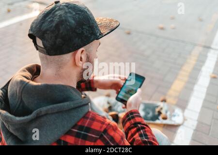 martelphone et communication. Un jeune homme en casquette, avec une barbe, utilise un smartphone. En arrière-plan se trouve une route et un skateboard. Vue arrière de dessus Banque D'Images