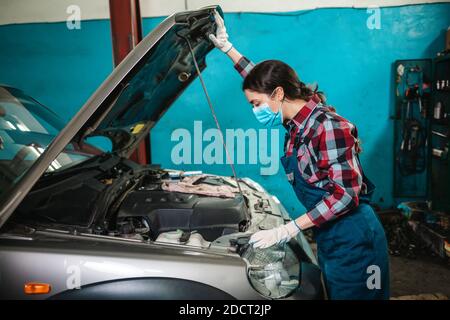 Portrait d'une jeune femme mécanicien en uniforme, portant des gants et un masque médical, ouvrant le capot d'une voiture. Vue latérale. Banque D'Images