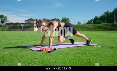 Jeune femme musclée portant un chapeau blanc faisant de l'exercice alpiniste sur le tapis en été ensoleillé. Magnifique entraînement de fille muscles de base sur le terrain de stade sur l'herbe verte fraîche. Concept d'entraînement. Banque D'Images