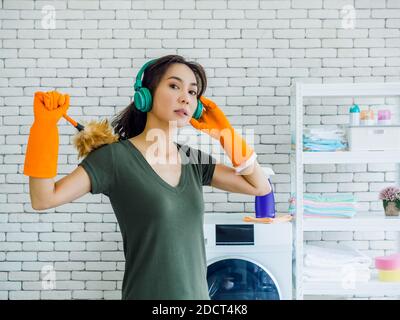 Belle jeune femme asiatique, femme au foyer portant des gants en caoutchouc orange, écoutant de la musique avec un casque vert et tenant un plumeau avec un visage fort Banque D'Images