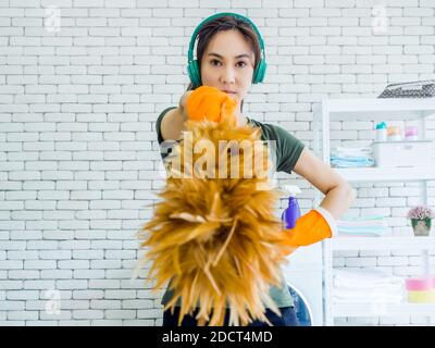 Belle jeune femme asiatique, femme au foyer portant des gants en caoutchouc orange, musique d'écoute avec casque vert et plumeau avec visage fort Banque D'Images