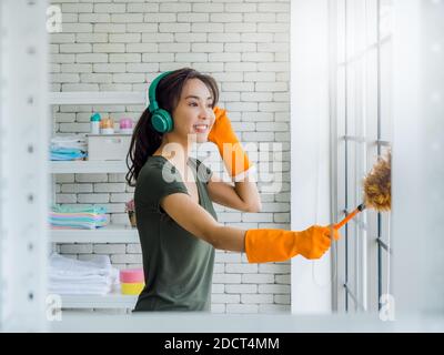 Belle jeune femme asiatique, femme au foyer portant des gants en caoutchouc orange, écoutant de la musique avec un casque vert et utilisant un plumeau nettoyant vent de verre Banque D'Images