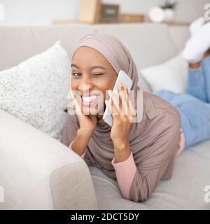 Magnifique femme noire dans hijab parlant sur téléphone mobile et elle se fait un clin d'œil sur son canapé à la maison Banque D'Images