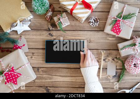 Maquette de tablette de bureau. Pose à plat de la main de la femme avec un smartphone, emballage cadeau de Noël. Présente sur fond de bois avec éléments de décoration, vue de dessus Banque D'Images