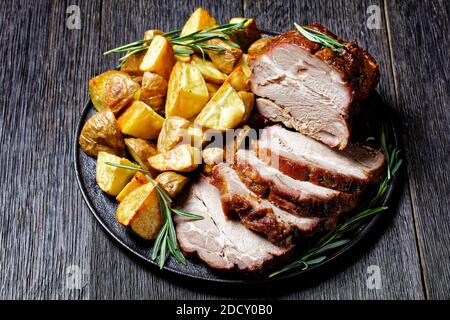 Filet de porc rôti entier servi avec des quartiers de pommes de terre cuits, romarin sur une assiette noire sur fond de bois foncé, vue du dessus, gros plan Banque D'Images
