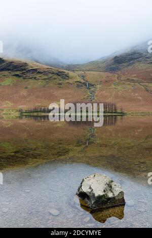 Réflexions de belles montagnes Cumbriennes dans le lac Buttermere avec une roche en premier plan sur un matin d'hiver brumeux. Banque D'Images