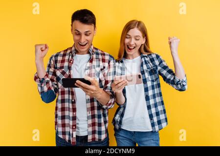 un couple heureux, un homme et une femme, jouer à des jeux vidéo sur un smartphone, se réjouir et montrer un geste gagnant, sur un fond jaune isolé Banque D'Images